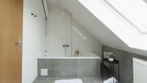 Une salle de bain moderne avec un puits de lumière, une baignoire, une douche et une petite plante posée sur une table d’appoint.