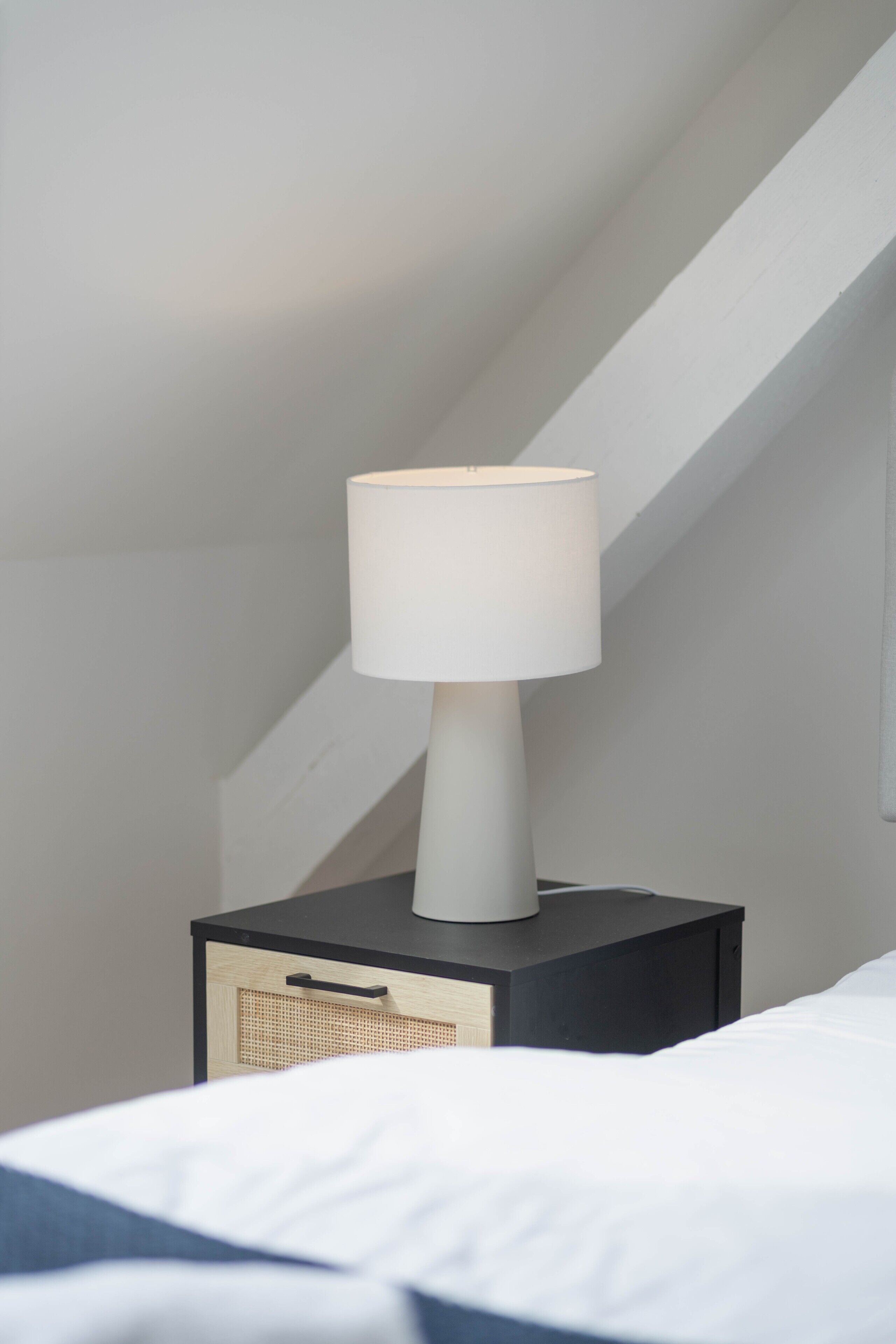 Una lampada da comodino bianca su un comodino nero con un cassetto, un letto con biancheria da letto bianca e un soffitto a falde.