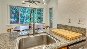 Cucina con piano di lavoro in granito, lavello in acciaio inossidabile e rubinetto moderno. Si nota un tavolo da pranzo in legno e sedie, oltre a un'ampia finestra con vista sugli alberi.
