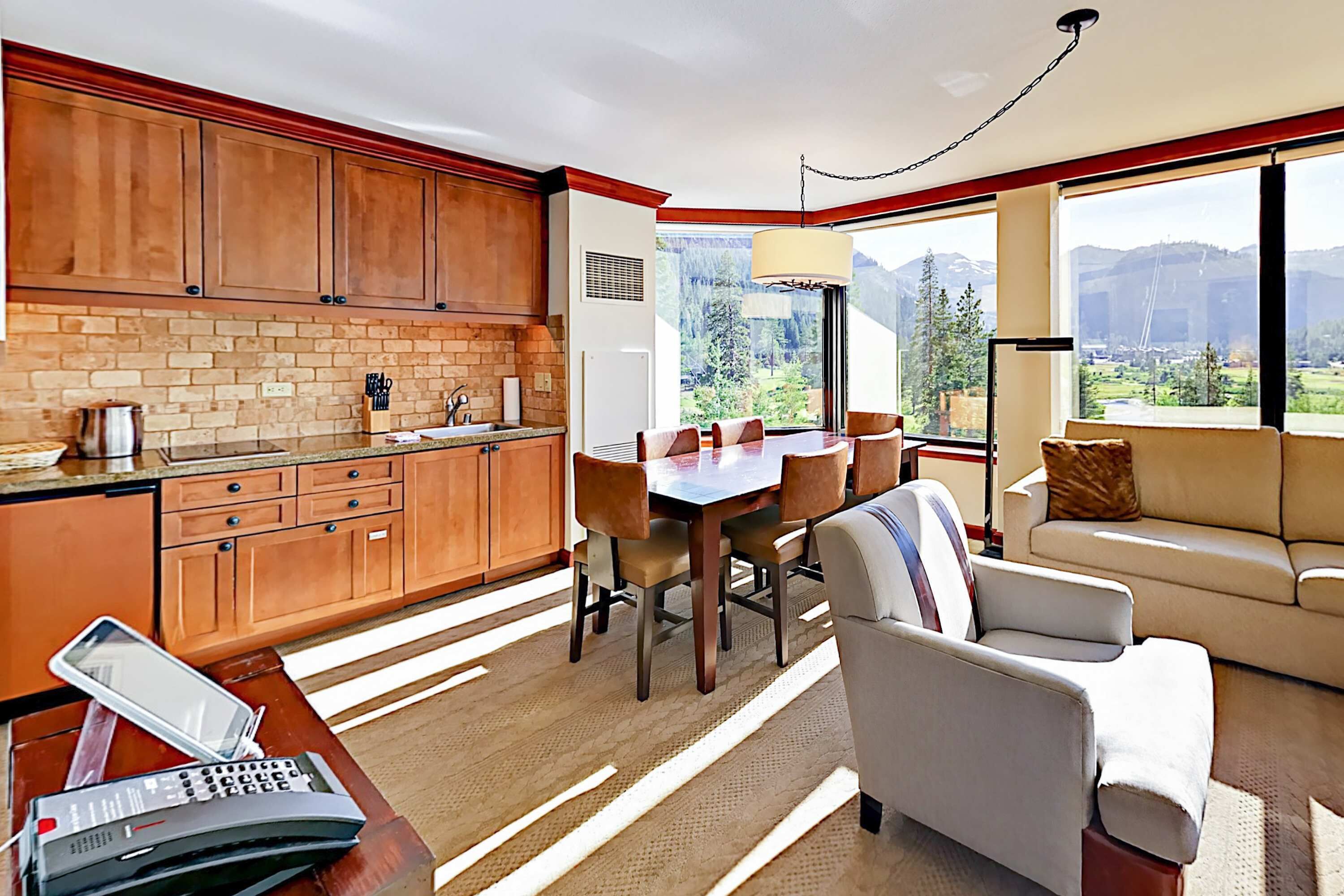 A modern kitchen with wooden cabinets, a dining area with a table and chairs, a sofa, and a large window with a mountain view.