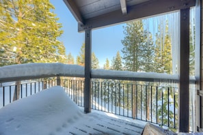 A snow-covered balcony with a metal railing, wooden beams, and a view of snow-laden trees. - Rustic-chic condo with pool, sauna, hot tub, tennis, fireplace, game room & deck (Truckee)