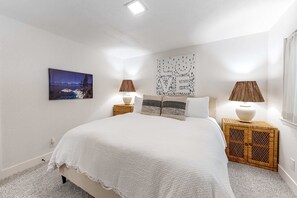 Un dormitorio con una cama grande, dos mesitas de noche, un buró, una lámpara de pared, un cuadro enmarcado y una ventana con cortinas.