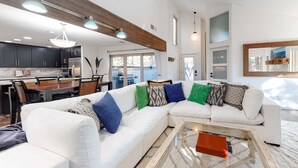 Ein modernes Wohnzimmer mit einem weißen Ecksofa, einem Glastisch und einem Essbereich mit dunklen Schränken und einem Holzbalken an der Decke.