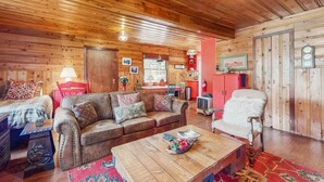 Ein gemütliches Blockhaus-Interieur mit Holzbalkendecke, einer Couch, einem Sessel, einem Esstisch und einer Küchenzeile.