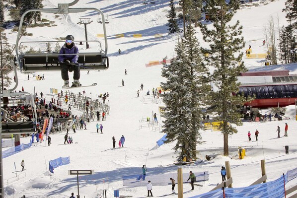 A ski lift with skiers descending, a ski slope with people, and a ski lodge in the background. - Condo with fireplace, balcony, washer&dryer, hot tub, & basketball & tennis courts (Truckee)