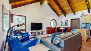 Ein modernes Wohnzimmer mit großem Fernseher, blauen Sofas, Holzboden und einer Küchenzeile.