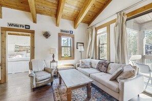 Une pièce chaleureuse avec un plafond en bois, un canapé, un fauteuil, une table et une fenêtre avec des rideaux.