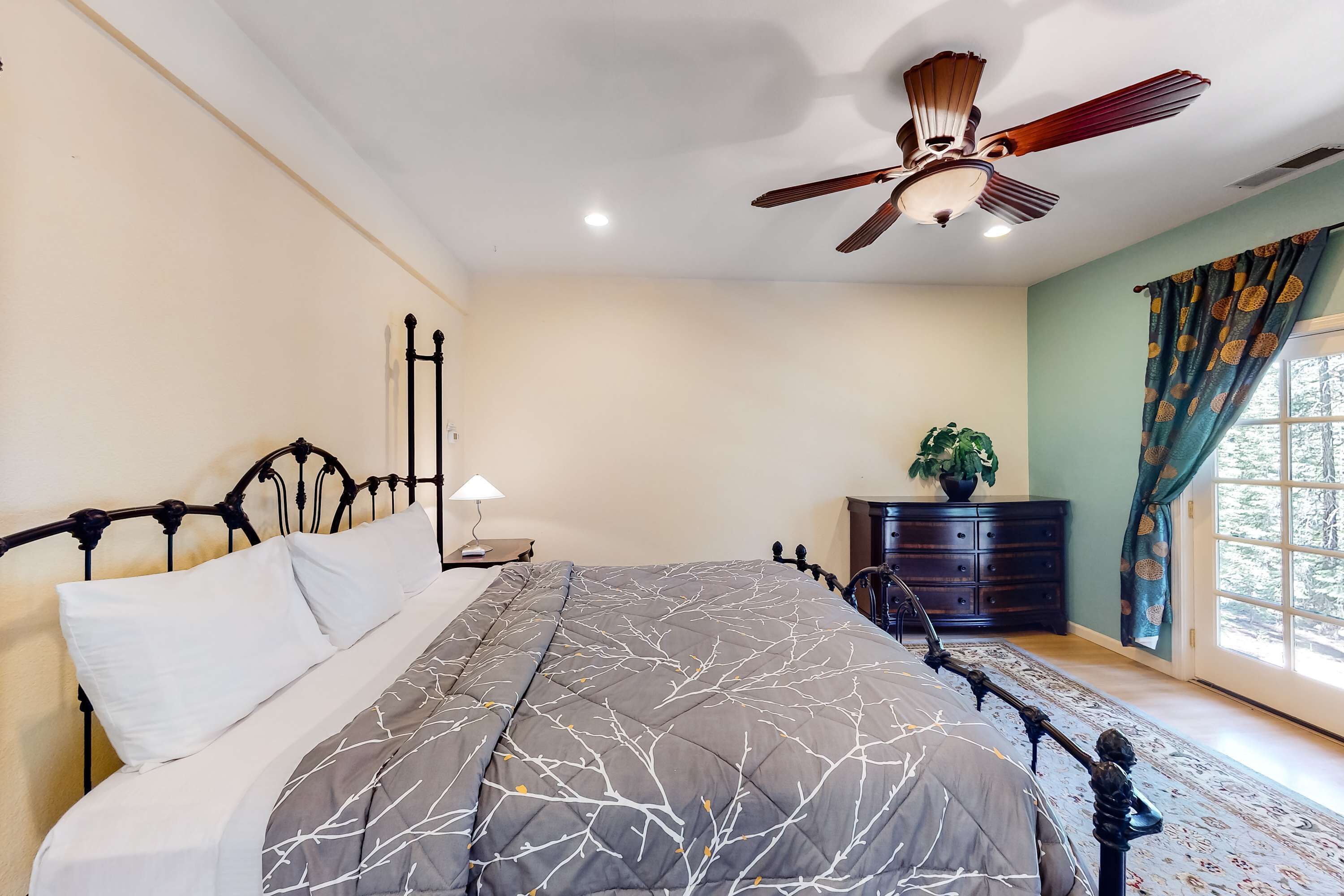 A bedroom with a bed, a ceiling fan, a dresser, a window with curtains, and a nightstand with a lamp.