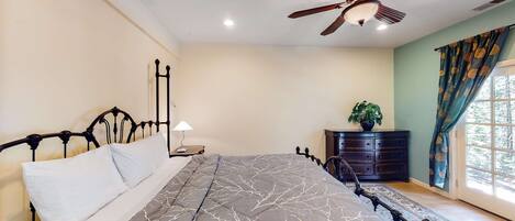 A bedroom with a bed, a ceiling fan, a dresser, a window with curtains, and a nightstand with a lamp.