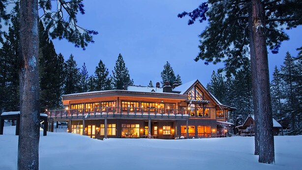 Un grand chalet à étages, doté d’un porche couvert et d’un balcon au deuxième étage, entouré de neige et d’arbres.