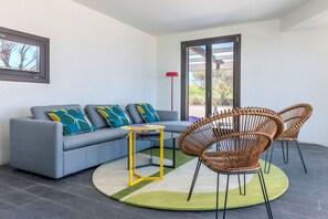 Een moderne woonkamer met een grijze bank, kleurrijke kussens, een kleine gele tafel, rieten stoelen en een groot raam met uitzicht.