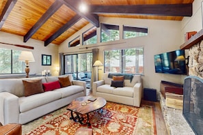 A spacious living room with a high ceiling, large windows, a fireplace, and a television.