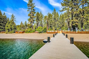 A wooden pier leading to a beach with clear water, surrounded by tall evergreen trees. - Pristine Modern Condo with Lake Views, Fireplace, Access to Private Beach (Tahoe City)