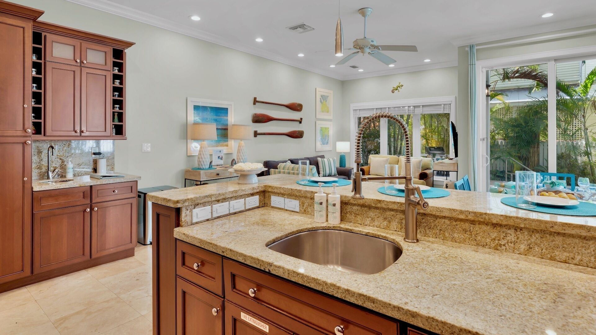 Una cocina con encimeras de granito, un lavamanos de acero inoxidable, armarios de madera y un comedor con vista al exterior.