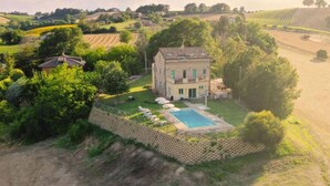 A large house with a swimming pool surrounded by a fence, set in a rural landscape with fields and trees. - Stunning private villa for 11 guests with WIFI, private pool, A/C, TV and patio (Montalto Delle Marche)