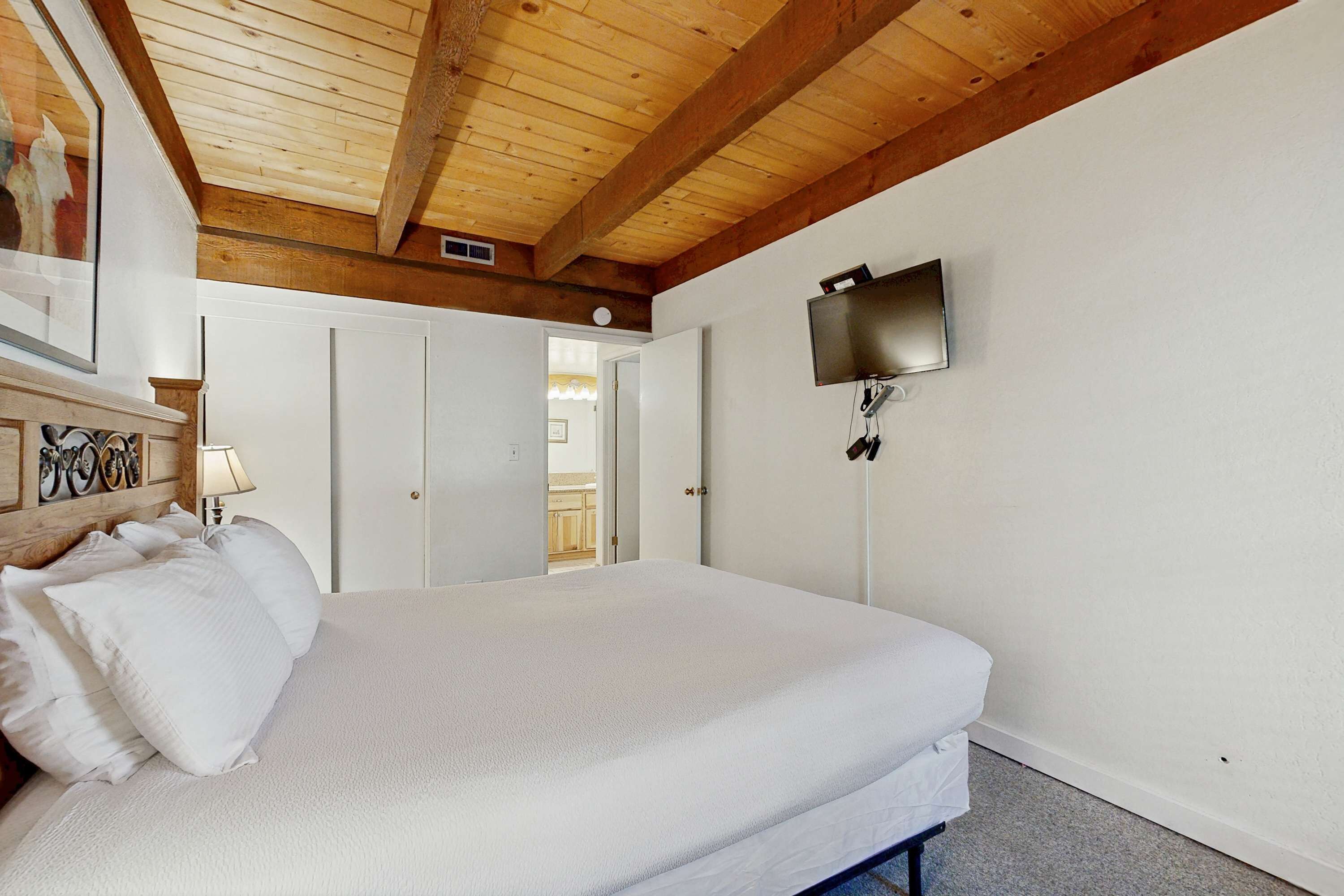 A bedroom with a wooden ceiling, a bed with white bedding, a TV mounted on the wall, and a door leading to another room.