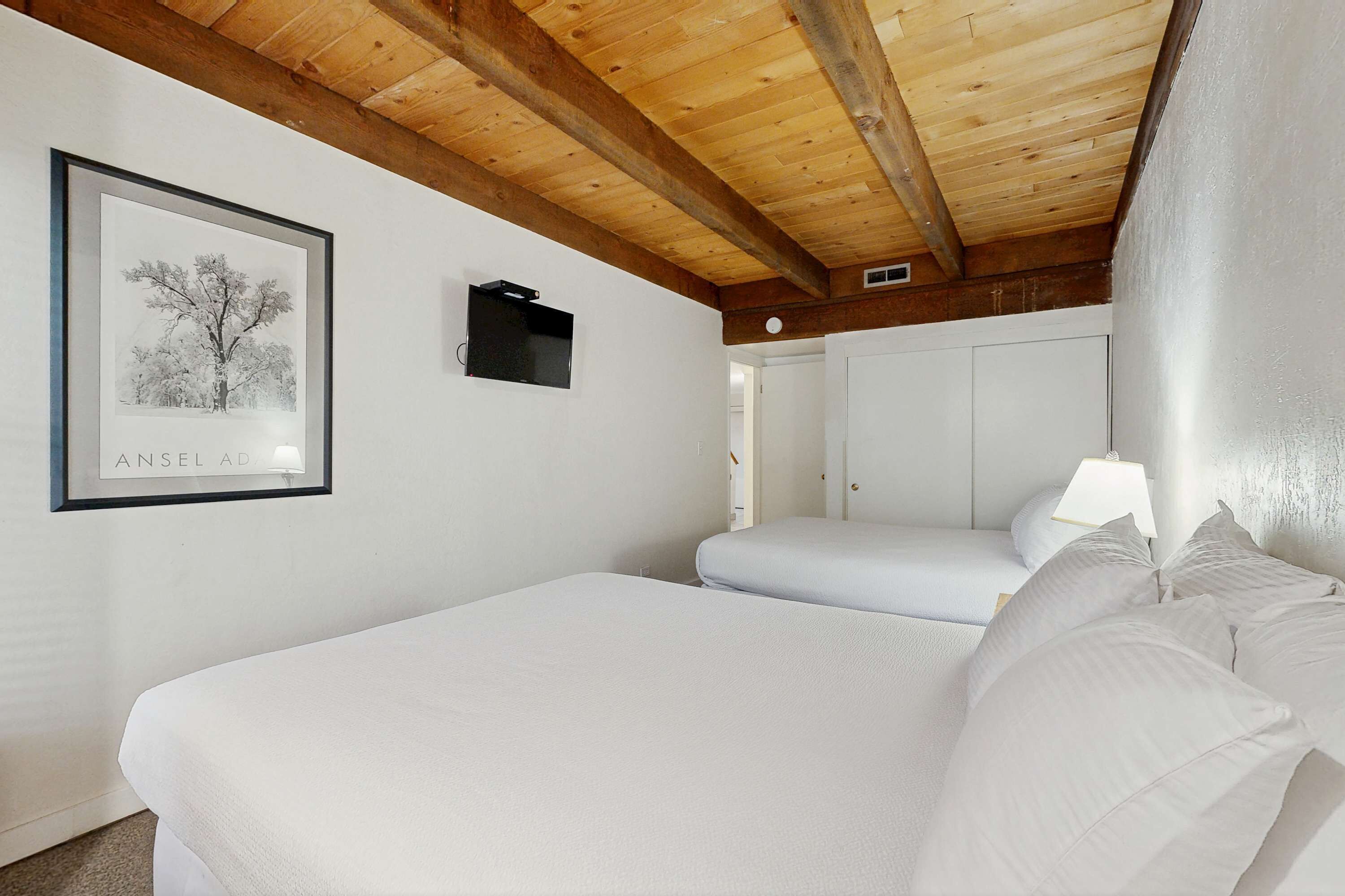 A bedroom with a wooden ceiling, a framed picture on the wall, a television, and a bed with white bedding.