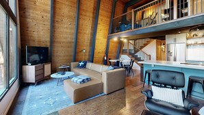 A spacious living area with a large sofa, a television, and a kitchen area with a bar. - Woodland home in quiet location with deck, fireplace & entertainment options (Kings Beach)