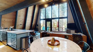 A modern kitchen with a central island, wooden beams, large windows, and a dining area with a round table. - Woodland home in quiet location with deck, fireplace & entertainment options (Kings Beach)