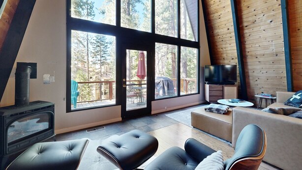 A modern living room with a fireplace, large windows, a TV, and a cozy seating area. - Woodland home in quiet location with deck, fireplace & entertainment options (Kings Beach)