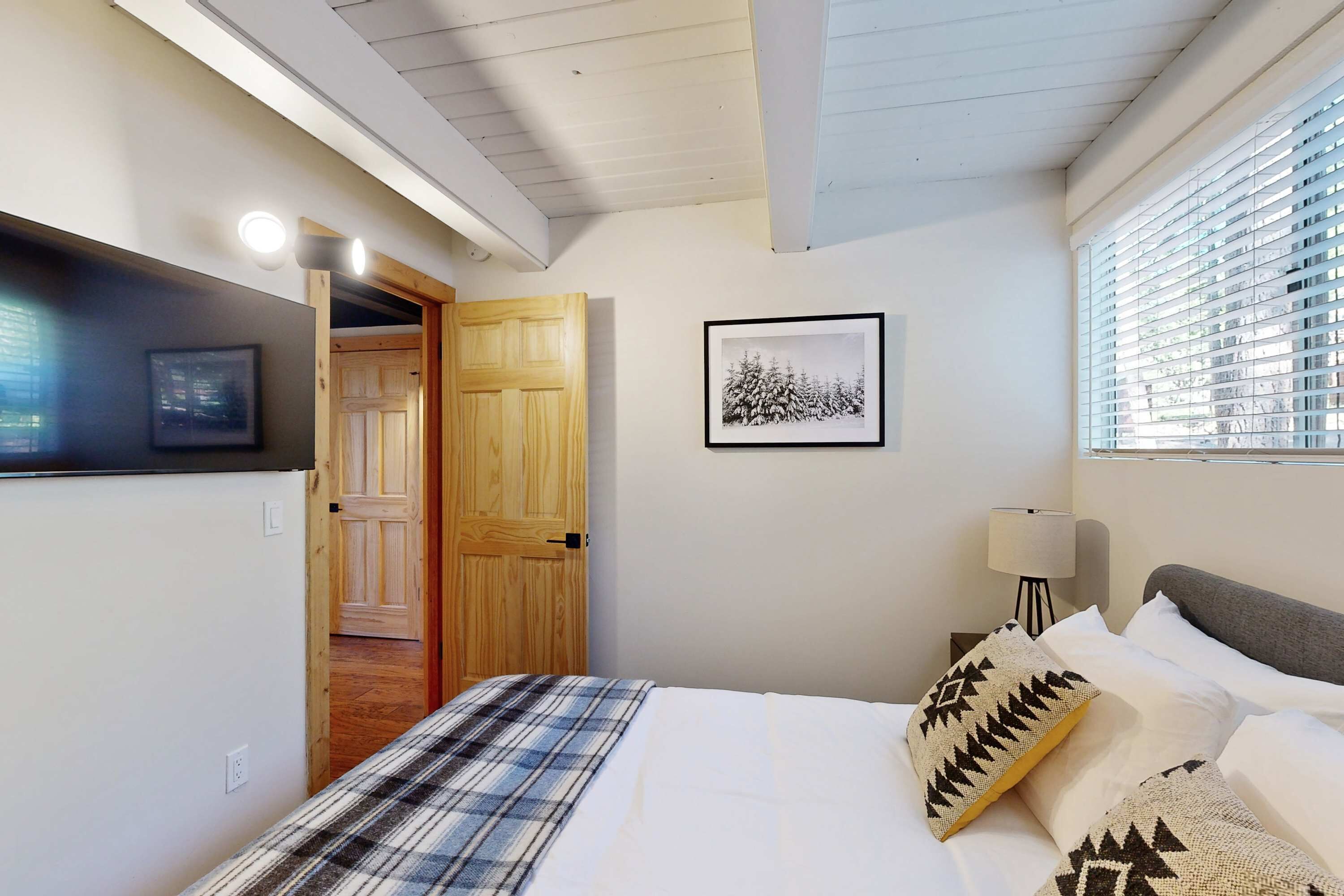 A bedroom with a bed, a wooden door, a framed picture, a lamp, and a window with blinds.