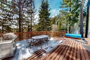 Eine Holzterrasse mit Grill, Tisch und Stühlen, umgeben von hohen Bäumen.