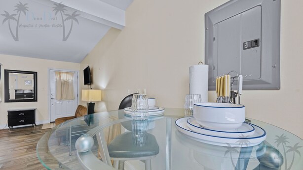 A modern dining area with a glass table, chairs, a white and blue tableware set, a wall-mounted TV, and a door with a towel. - Oak Shores Studio 13 | Just a Short Walk to the Beach! (Biloxi)