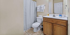 A bathroom with a toilet, a shower curtain, a sink with toiletries, and a wooden cabinet. - Oak Shores Studio 13 | Just a Short Walk to the Beach! (Biloxi)