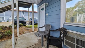 A covered porch with two chairs, a door, and a window overlooking a parking area. - Oak Shores Studio 13 | Just a Short Walk to the Beach! (Biloxi)
