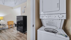 A laundry area with a washing machine, a black cabinet, and a wooden stool. - Oak Shores Studio 13 | Just a Short Walk to the Beach! (Biloxi)