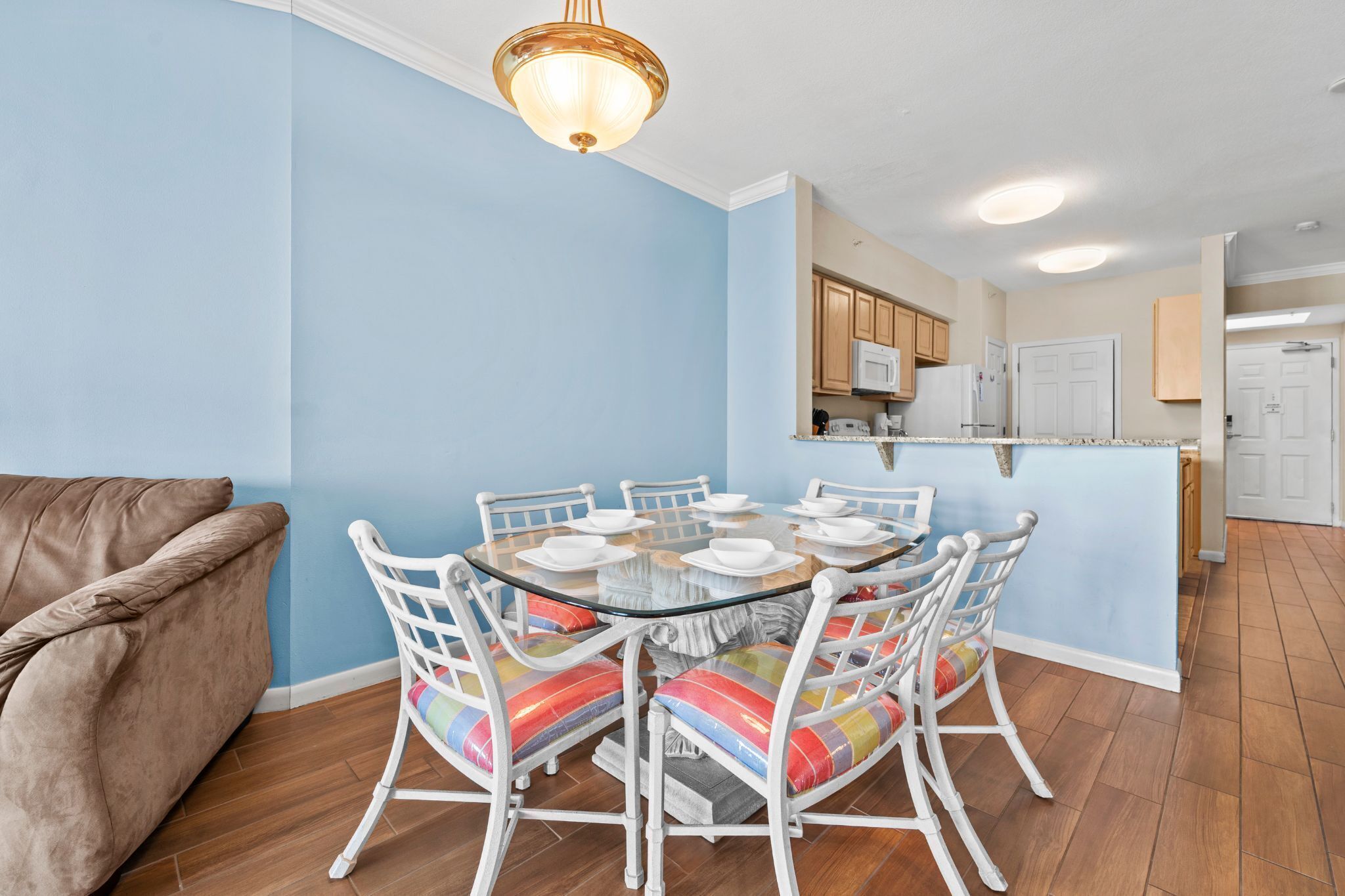 A dining area with a round glass table, white chairs with colorful cushions, a brown leather couch, and a kitchen in the background.