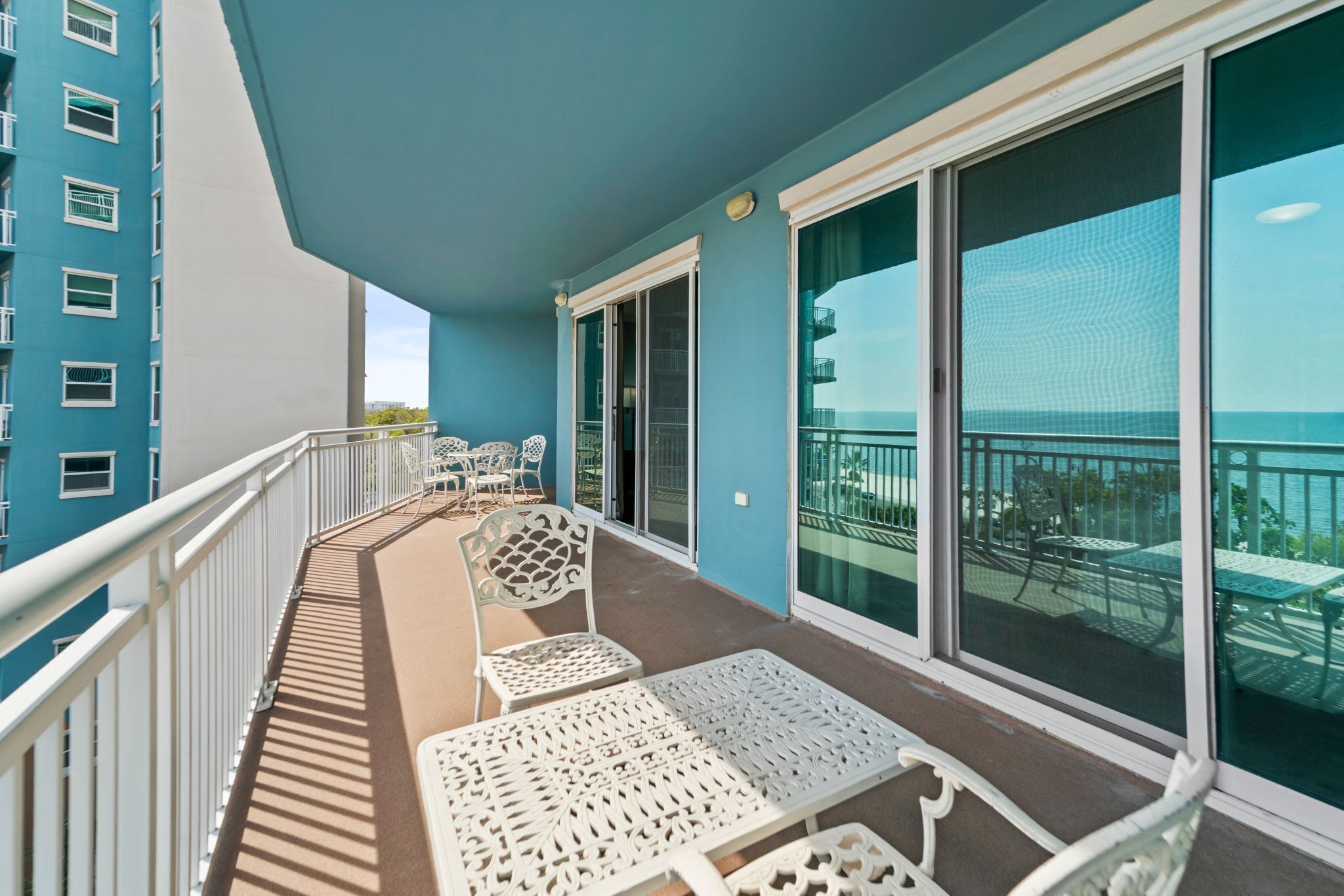 A balcony with white metal chairs and tables, a blue building, and a view of the sea.