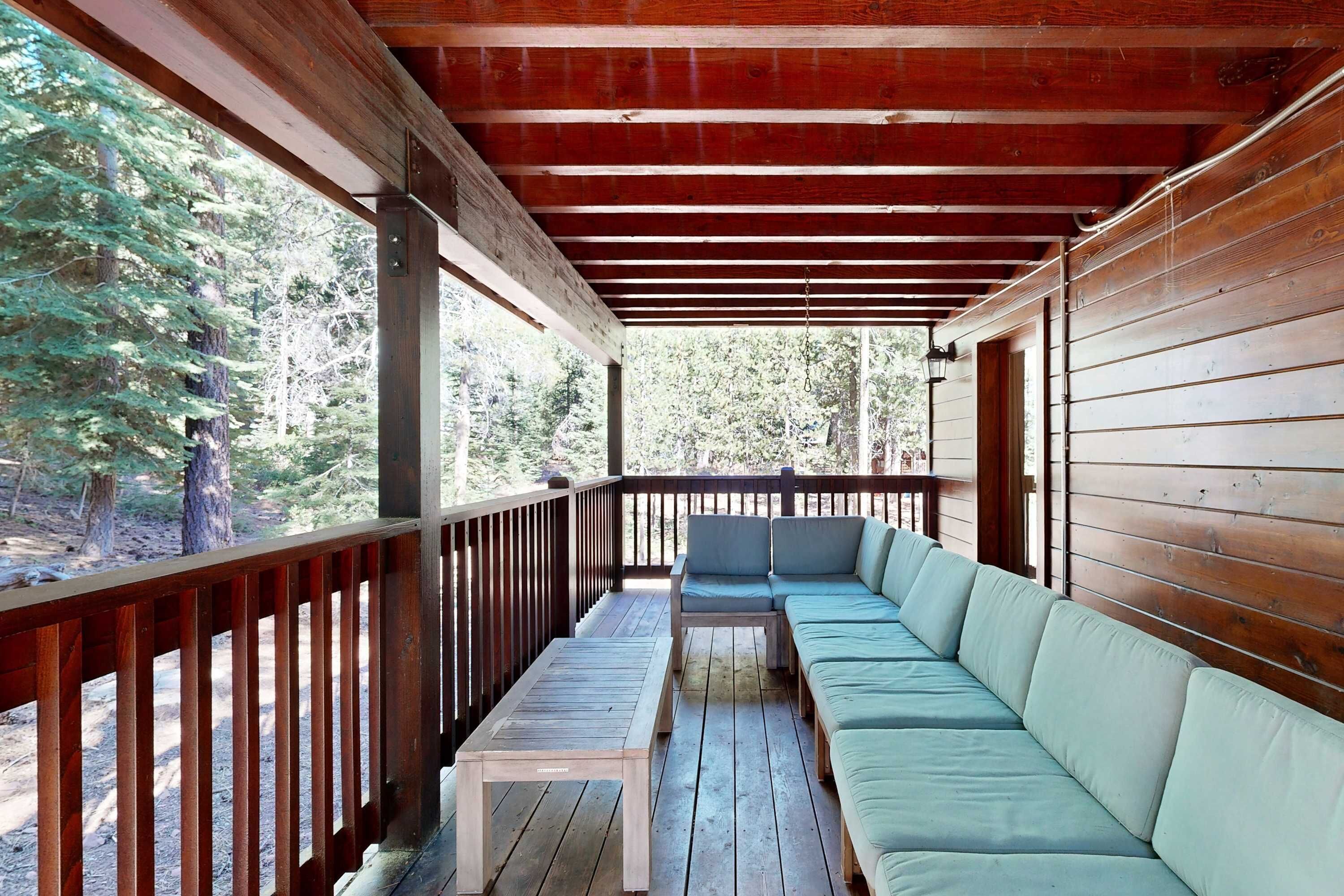 A covered deck with a wooden bench and a sofa, surrounded by a wooden railing, overlooking a forested area.
