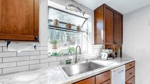 A kitchen with a stainless steel sink, wooden cabinets, a window with a view of trees, and a dish rack with dishes. - Ski getaway with amazing mountain views, fireplace, pool, & river path access (Olympic Valley)