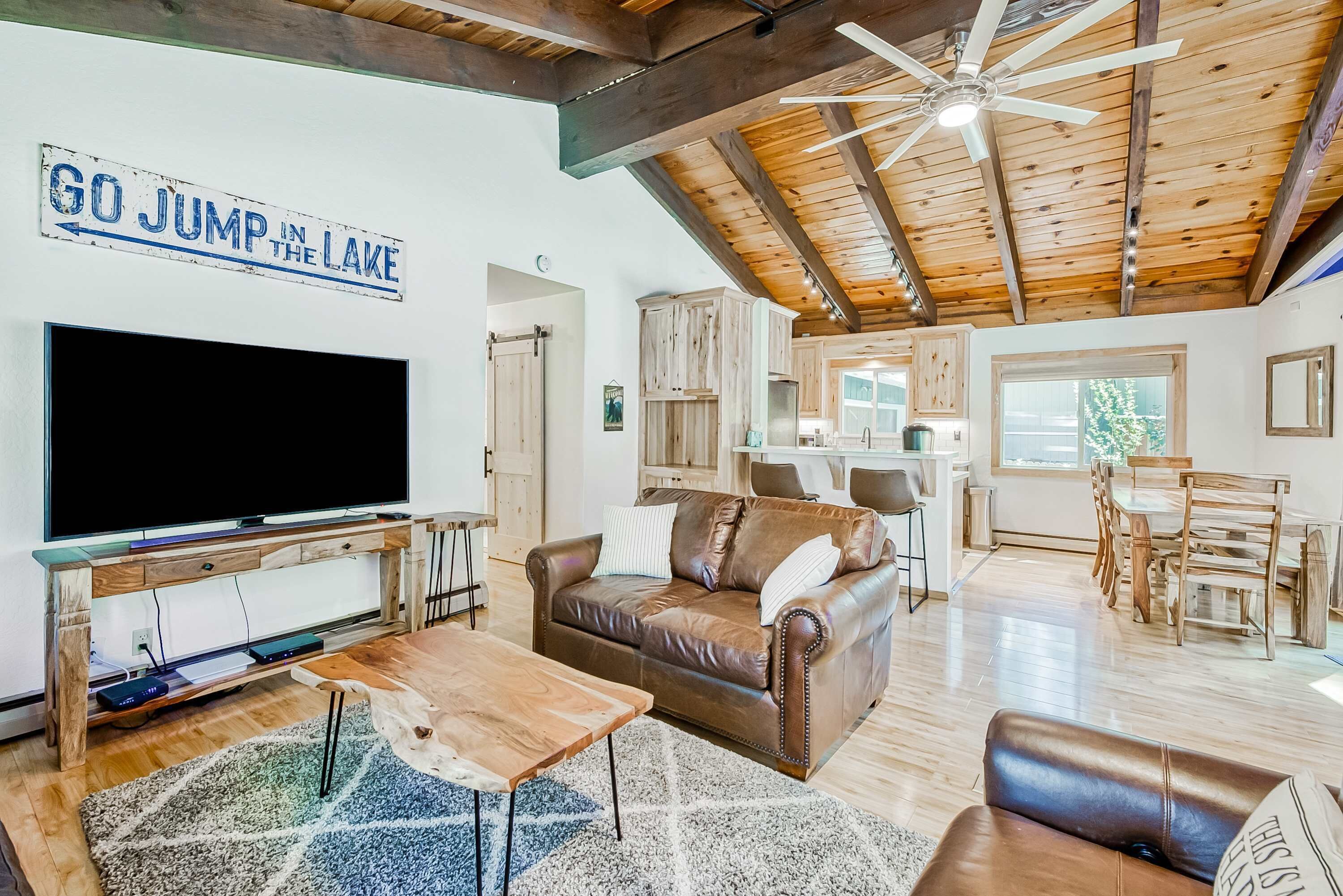 A spacious living room with a large flat-screen TV, a brown leather sofa, a wooden coffee table, and a ceiling fan.