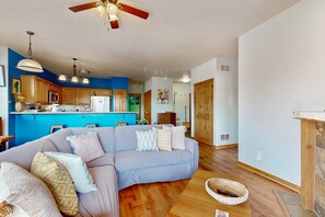 Ein Wohnzimmer mit einer Couch, Kissen, einem Deckenventilator, Pendelleuchten und einer Küche im Hintergrund.