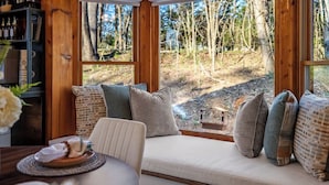 Ein gemütlicher Sitzbereich mit gepolstertem Sitz, einem Esstisch mit Unterlage und Blick auf ein bewaldetes Gebiet durch große Fenster.