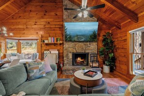 A cozy cabin interior with a stone fireplace, wooden walls, a ceiling fan, and a view of the outdoors through large windows.