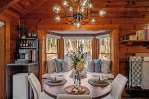 A wooden cabin interior with a dining table, chairs, a chandelier, and a view through large windows.