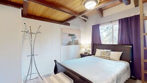 Et soveværelse med træloft, en seng med hvidt sengetæppe, et natbord med lampe og et vindue med lilla gardiner.