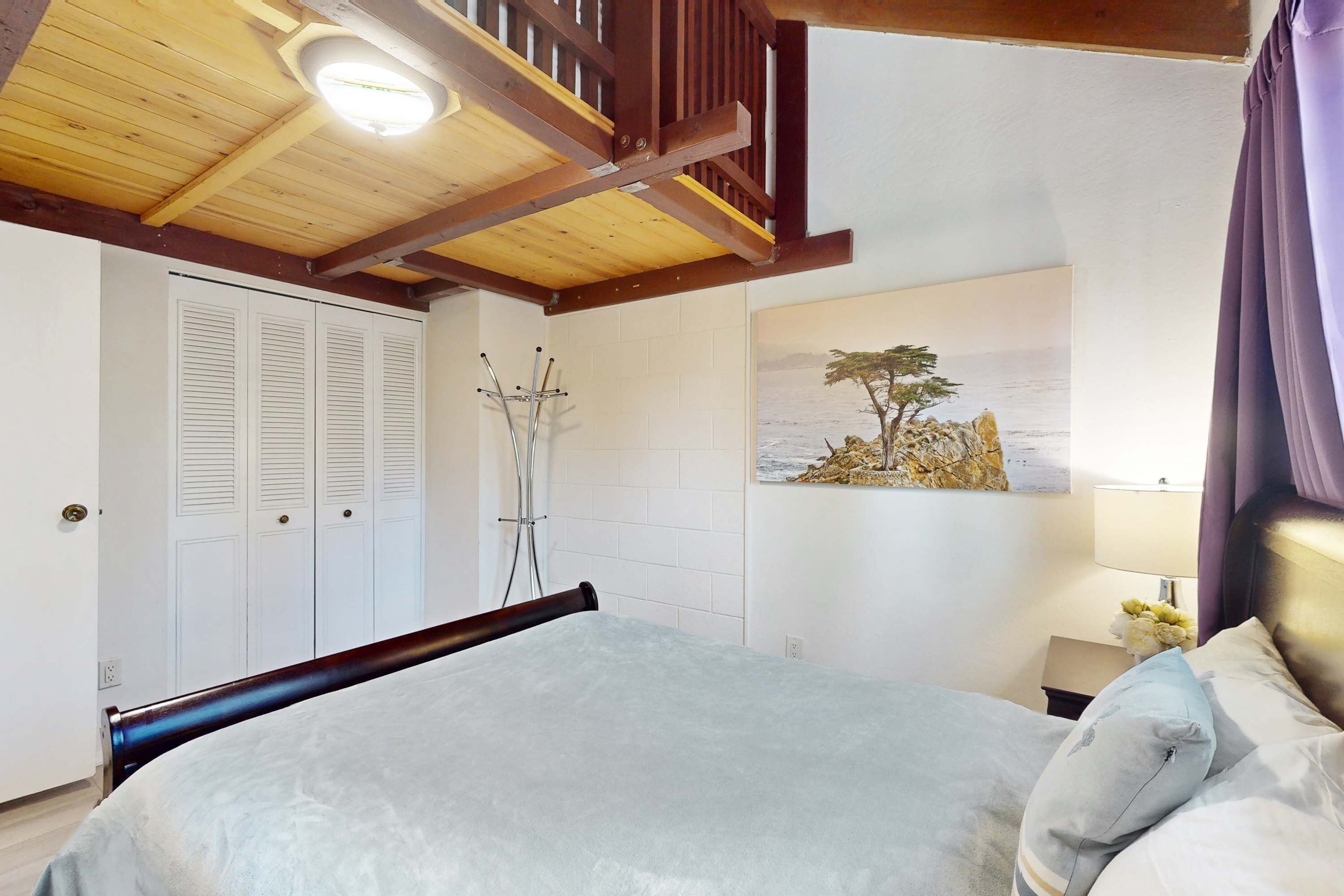 A bedroom with a bed, wooden ceiling beams, white walls, a painting, and a nightstand with a lamp.
