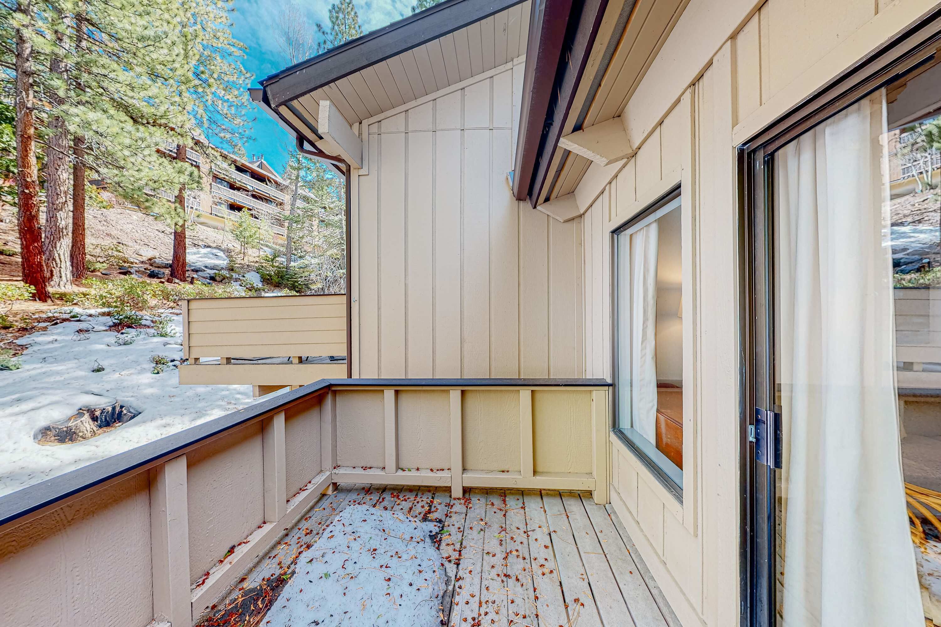 Un espace extérieur abrité doté d’une terrasse en bois, d’une porte-fenêtre coulissante et offrant une vue sur des arbres et un bâtiment en arrière-plan.