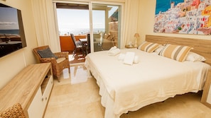 Habitación de hotel con cama, una silla, una mesa, televisor y un cuadro de un pueblo costero.