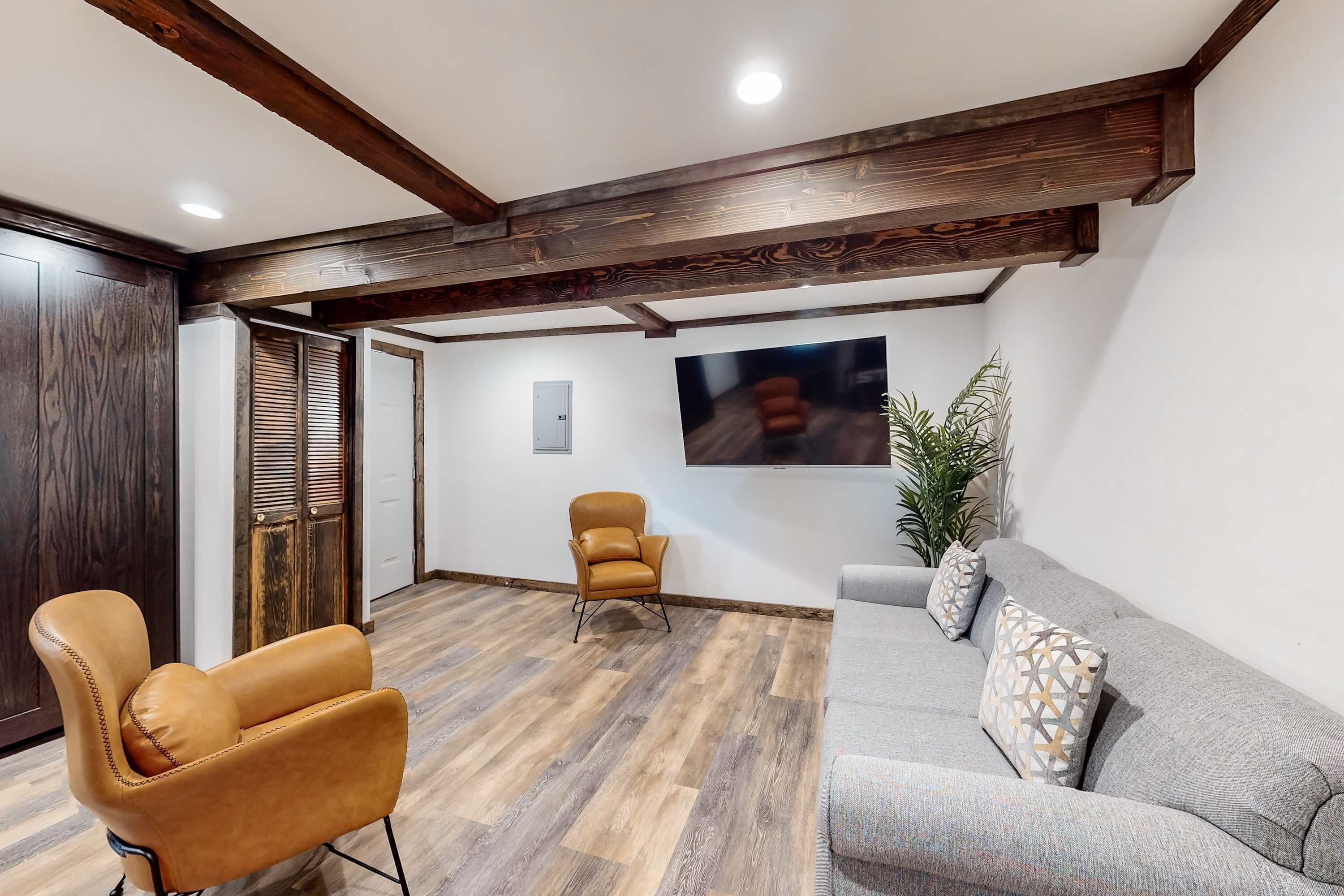 Une pièce avec un plafond en bois, un canapé gris, un fauteuil en cuir marron, un téléviseur fixé au mur et une plante.