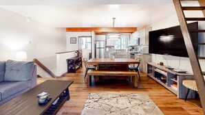 Ampla sala de estar com mesa de jantar de madeira, sofá, televisão e cozinha ao fundo.