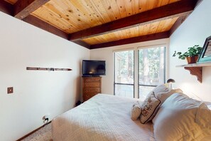 Ein Schlafzimmer mit Holzboden, einem Fernseher auf einer Kommode, einem Bett mit Kissen und einem Fenster mit Jalousien.