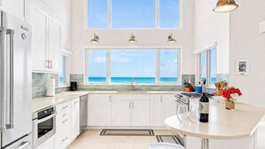 Una cocina moderna con armarios blancos, una isla central, vista al océano y un comedor con mesa y sillas.