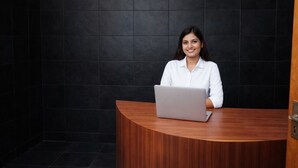 Une personne est assise à un bureau en bois, devant un ordinateur portable, dans une pièce dont les murs sont recouverts de carreaux sombres.