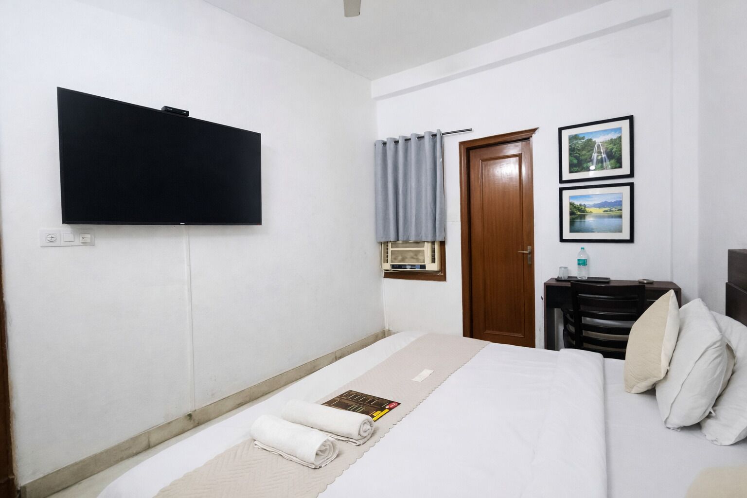 Habitación de hotel con cama, televisor de pantalla plana, puerta de madera y dos cuadros en la pared.