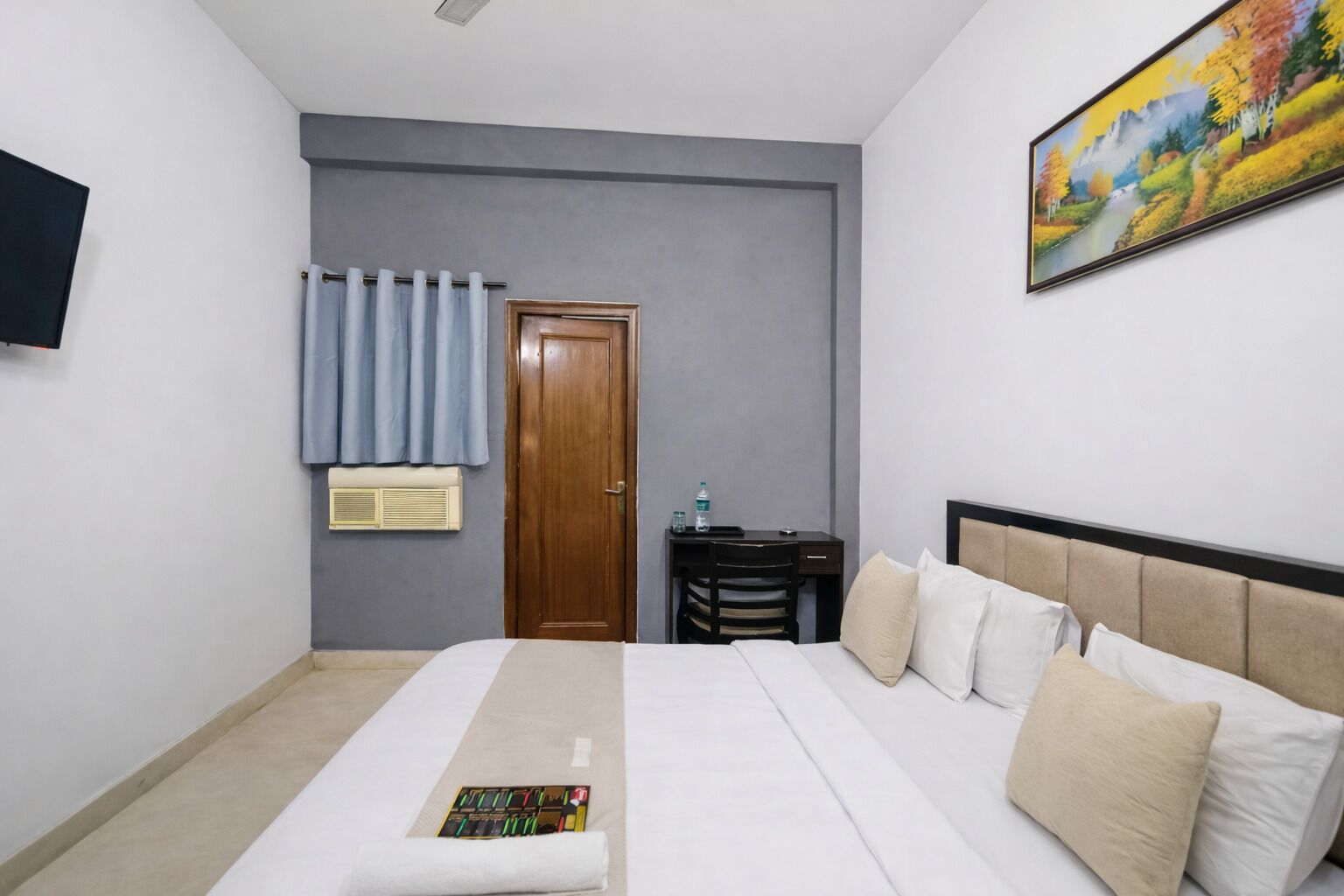 A hotel room with a bed, a desk, a chair, a door, a painting, and a TV on the wall.